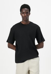 T-shirt noir en tricot côtelé à manches courtes, décolleté rond et coupe décontractée, associé à un pantalon de couleur claire.
