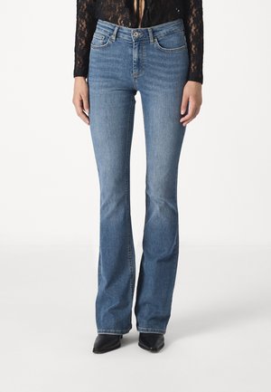 Femme portant un jean bootcut bleu clair avec bouton et fermeture éclair à l'avant, associé à des chaussures noires pointues et un haut en dentelle noire à manches.