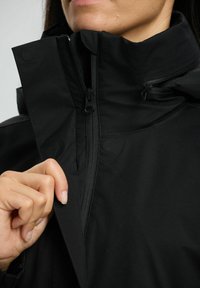 Dłoń osoby regulująca zamek błyskawiczny i klapę czarnej kurtki z wysokim kołnierzem, ukazująca strukturę materiału i detale zapięcia.