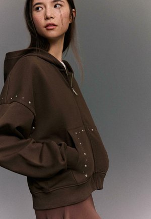 Young woman wearing oversized brown hoodie with silver studs near zipper and pockets, looking to the side against plain gray background.