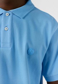 Polo shirt azzurro chiaro in cotone testurizzato, con colletto a tre bottoni e un logo circolare blu sul lato sinistro del petto.