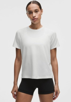 Kvinde med trukket hår iført en enkel hvid kortærmet top og sorte tætsiddende shorts, stående mod en ensfarvet baggrund.