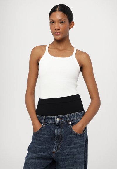 Top in cotone bianco con spalline sottili e una cintura a coste nera, abbinato a jeans a vita alta blu scuro con tasche laterali.