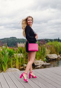 Blondýnka v černých šatech jde po dřevěném molu u vody, má na sobě růžové vysoké podpatky a nese růžovou kabelku s bílými puntíky.