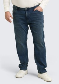 Blaue Jeans aus Denim mit geradem Bein, fünf Taschen und vorderem Knopfverschluss, mit subtiler Ausbleichung und kontrastierenden Nähten. Tragen mit weißen Schuhen.