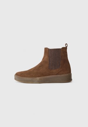 Brown suede ankle boots with elastic side panels and a textured fabric insert. Flat rubber sole, stitched detailing, and a pull tab.