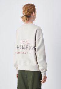 Felpa grigia chiara con una vestibilità ampia, caratterizzata da una scritta "CHAMPION" sbiadita in nero e rosso e una grafica con un numero all'interno di un riquadro sul retro.