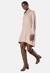 Abito beige oversize con scollo a cowl e lunghe maniche a sbuffo, abbinato a stivali alti fino al ginocchio con tacco a blocco marroni. Tessuto morbido.