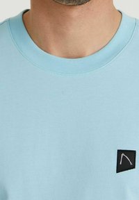 Jasnoniebieska koszulka z krótkim rękawem i okrągłym dekoltem. Posiada czarną naszywkę z białym geometrycznym wzorem po lewej stronie klatki piersiowej.