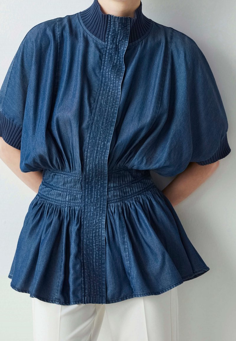 Denim peplumtop med hög ribbad krage, insydd midja och korta ärmar. Tyget är djupt blått med en slät yta.