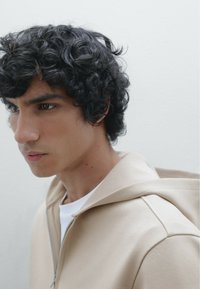 Haut à capuche beige zippé avec une finition texturée. Porté sur une chemise blanche, avec une capuche à cordon de serrage et une coupe décontractée. Cheveux foncés bouclés visibles.