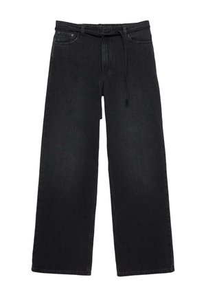 Femme portant un haut blanc sans manches et un pantalon noir à jambes larges, les mains dans les poches, debout devant un fond clair uni.