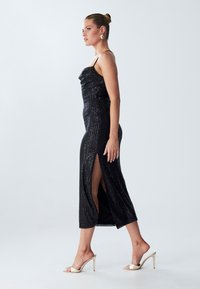 Abito da sera nero in paillettes con scollatura a cappuccio e spacco alto alla coscia. Vestibilità slim, lunghezza al polpaccio, sottili spalline a spaghetti e texture liscia.