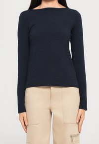 Femme portant un haut bleu marine moulant à manches longues avec un col bateau et un pantalon beige avec de grandes poches et un détail zippé.