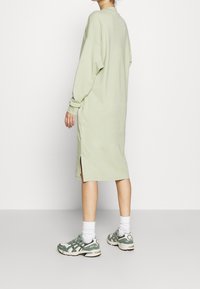 Olive green oversize sukienka z długim rękawem, bocznymi rozcięciami i luźnym krojem, zestawiona z szaro-zielonymi trampkami i białymi skarpetkami.