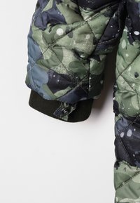 Manche de veste matelassée avec un motif camouflage vert, noir et bleu, dotée d'un poignet en ribbed noir et d'une patte à bouton pression.