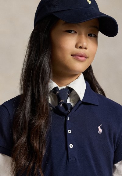 Young person with long dark hair wearing a navy cap, navy polo shirt with a pink logo, white collared shirt, and navy tie with small patterns.