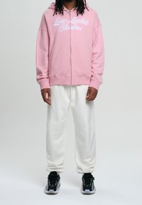 Sweat à capuche rose zippé avec texte brodé blanc, assorti d'un pantalon de survêtement blanc et de baskets noires. Les matériaux semblent doux et décontractés.