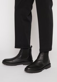 Bottes Chelsea noires en cuir avec des panneaux latéraux en élastique portées avec un pantalon noir à ourlet sur un fond clair.