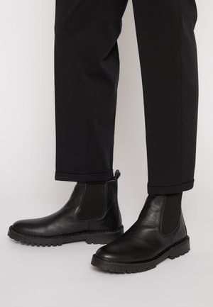 Zwarte leren Chelsea boots met elastische zijpanelen, gedragen met zwarte omgeslagen broeken op een lichte achtergrond.