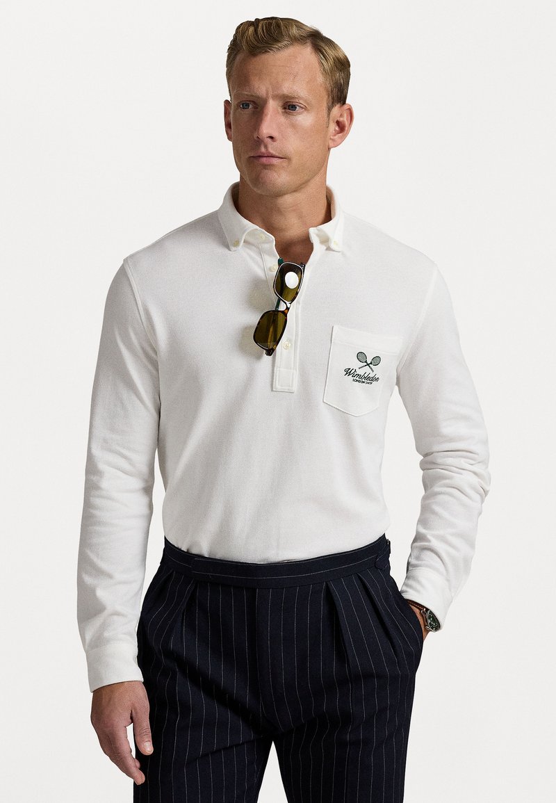 Cămașă polo albă cu mâneci lungi, guler cu nasturi și textură tricotată, având un buzunar cu un logo de rachetă de tenis verde. Asortată cu pantaloni cu dungi fine.