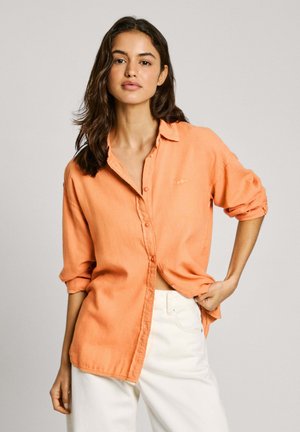 Camicia arancione a maniche lunghe con maniche arrotolate, realizzata in tessuto leggero; presenta una tasca e una vestibilità comoda, abbinata a pantaloni bianchi.