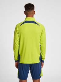 Langærmet pullover med høj krave i lys neongrøn, med marineblå detaljer og åndbart materiale. Sammen med marineblå shorts.