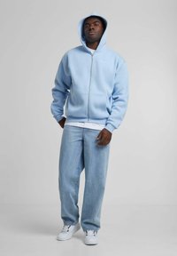 Lyseblå zip-up hettegenser laget av myk stoff med en frontlomme. Kombinert med løstsittende lyse denimjeans og hvite joggesko.