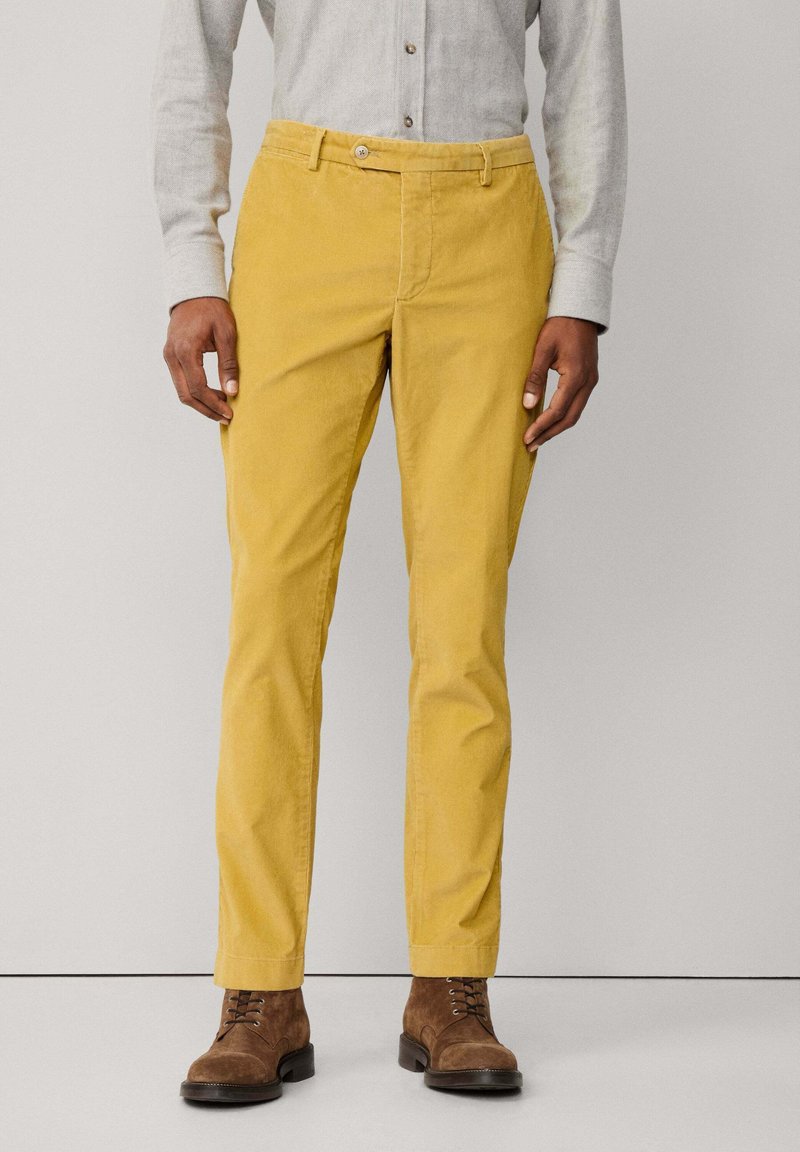 Keltaiset korduroy-housut, joissa on slim fit -malli, vyötärö, jossa on napin kiinnitys, ja etutaskut, yhdistettynä ruskeisiin mokkasaappaisiin.