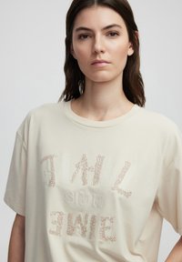 Camiseta de algodón beige con manga corta, que presenta letras texturizadas y adornos. Diseño de cuello redondo, corte relajado.
