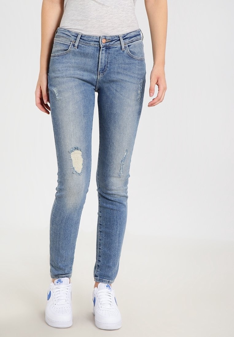 Jeans de mezclilla azul ajustados con parches desgastados, una cintura de media altura y un ligero desvanecimiento. Combinados con zapatillas blancas.