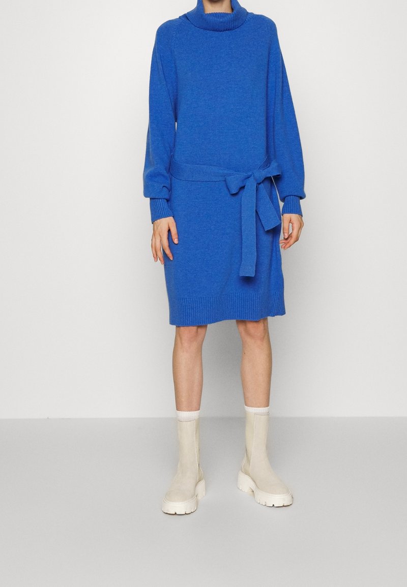 Abito blu a maglia con colletto alto, vita cintrata e polsini a costine. Abbinato a stivaletti beige alla caviglia. Texture liscia e vestibilità comoda.