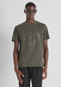 Khaki t-shirt van katoen met korte mouwen, met een tijgerprint in een uitgesneden stijl op de voorkant. Gecombineerd met donkere broeken.
