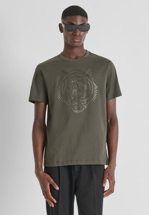 Camiseta caqui de algodón de manga corta, con un diseño gráfico de un tigre en estilo recortado en la parte delantera. Combinada con pantalones oscuros.