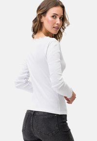 Top bianco a maniche lunghe con texture a costine e scollatura ondulata. Indossato con jeans scuri, evidenziando una forma aderente.