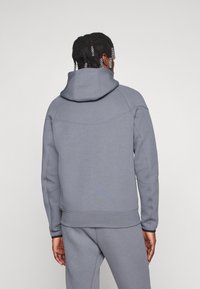 Felpa con cappuccio in grigio chiaro, realizzata in tessuto morbido, con yoke cucito e polsini a coste. La vista posteriore mostra una vestibilità rilassata e un design senza cuciture.