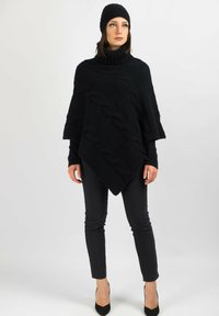 Svart stickad poncho med ett panel av kabelmönster, med hög krage. Matchad med svarta slim-fit byxor och klackar. Svart beanie fulländar looken.