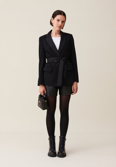 Blazer nero su misura con cintura, abbinato a pantaloni corti in pelle, collant neri e stivali alla caviglia con lacci. Piccola borsa nera con dettagli in oro.