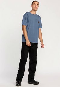 Blaues Kurzarm-T-Shirt mit einer Taschenverzierung, kombiniert mit schwarzen Cargohosen. Das Modell steht und trägt schwarze Schuhe. Einfaches, lässiges Outfit.
