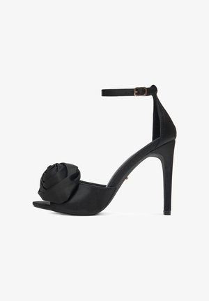 Czarne sandały na wysokim obcasie z zapięciem na kostkę i dużym detalem z tkaniny w kształcie róży z przodu, na otwartym palcu.