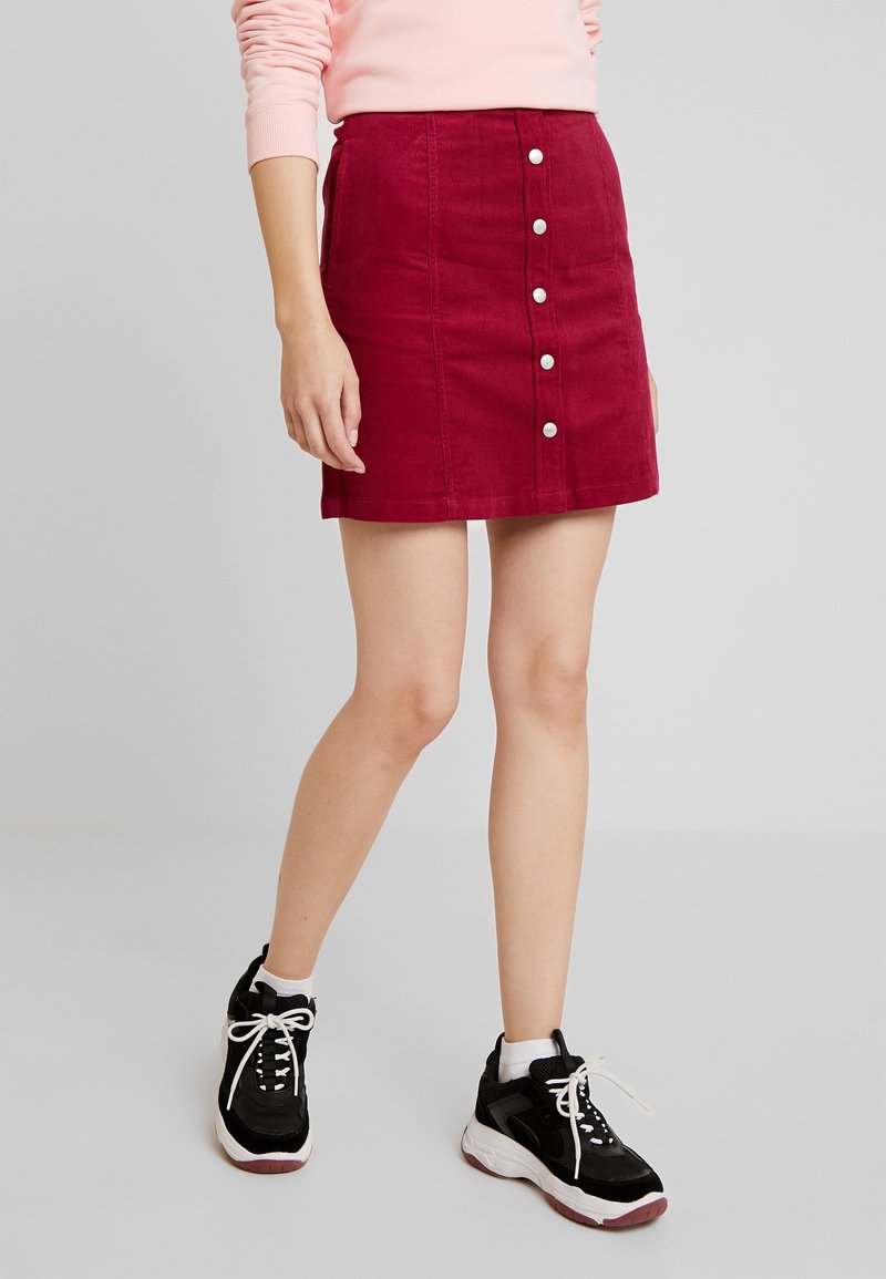 Červená manšestrová sukně se zapínáním na knoflíky vpředu a bočními kapsami, doplněná černobílými teniskami, na světlém pozadí.