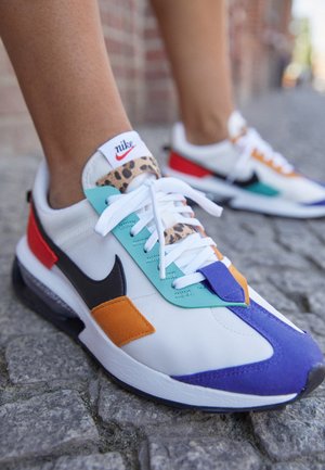 Baskets blanches Nike avec des accents multicolores incluant bleu, orange, sarcelle, motif léopard et rouge portées sur un trottoir pavé.
