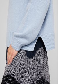 Pull en tricot bleu clair avec des poignets et un ourlet côtelés, présentant des fentes latérales. Porté par-dessus une jupe noire et blanche à motifs avec des designs géométriques.