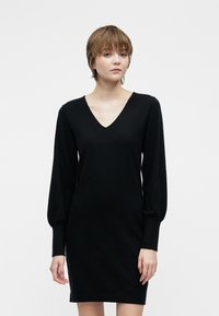 Mladý človek s krátkymi svetlohnedými vlasmi, oblečený v čiernych šatách s dlhým rukávom a výstrihom do V, stojí proti čistému bielému pozadiu.