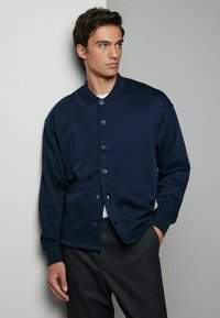 Cardigan bleumarin cu nasturi, cu guler, croi lejer, nasturi în față și manșete cu zimți. Purtați peste un tricou alb și asortați cu pantaloni negri.