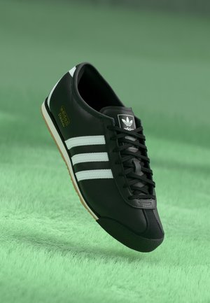 Sort Adidas sneakers med hvide striber og gummisål svævende over en grøn tekstureret overflade.