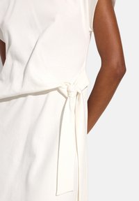 Jasno beżowa sukienka z krótkimi rękawami, z bocznym węzłem w pasie i gładkim, teksturowanym materiałem. Prosty i minimalistyczny design.