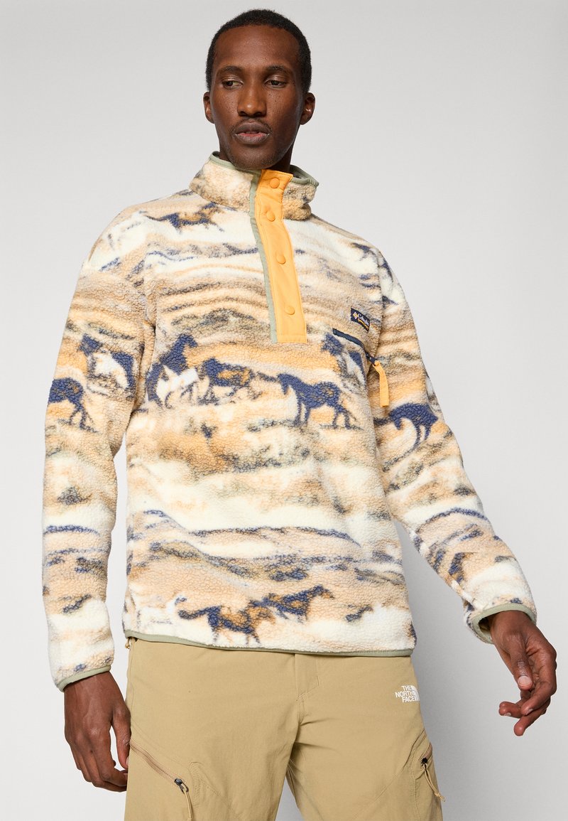 Pullover in pile beige con motivo di cavalli blu e marroni, colletto a metà zip e bottoni arancioni. Presenta una tasca sul petto sinistro con un tirante arancione.