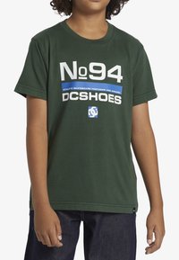 Zelené bavlněné tričko s kulatým výstřihem a krátkými rukávy, na přední straně s výrazným bílým a modrým grafickým textem "No 94 DC Shoes".