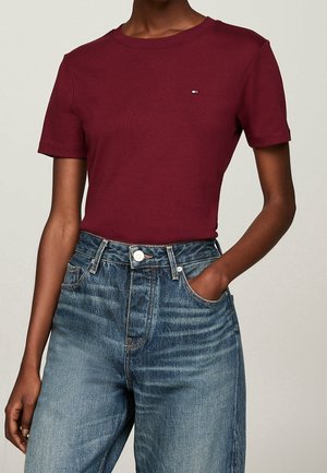 Mujer con camiseta ajustada de manga corta color burdeos con un pequeño logo y vaqueros azules de talle alto, con una mano en el bolsillo.